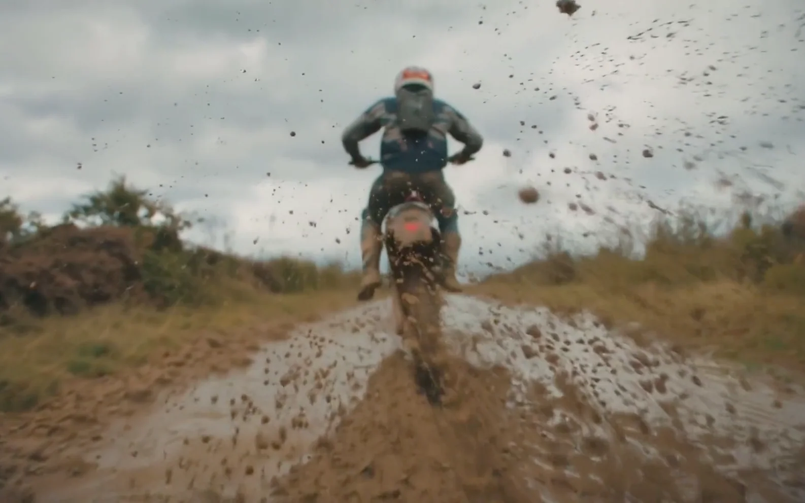 Rear tracking view of a motocross rider throwing mud toward the camera on a flooded off-road trail.